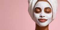 Young African American woman with white facial mask and towel on head relaxing during spa treatment on pink background for skincare routine.
