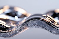 Macro shot of liquid metal wave with silver reflective surface and water drops creating dynamic flowing pattern against light gray background.