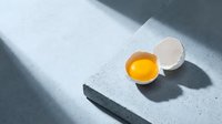 Cracked egg with bright yellow yolk on white speckled cutting board with dramatic shadows, kitchen cooking ingredient for breakfast recipes.