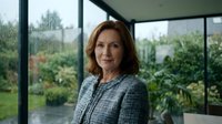 Mature woman with shoulder-length auburn hair and fair complexion wearing gray tweed blazer standing indoors near large windows overlooking garden. Professional portrait.