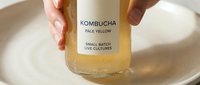 Hands holding glass bottle of pale yellow kombucha over ceramic bowl, showcasing fermented probiotic drink with small batch live cultures label for healthy lifestyle.
