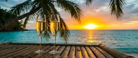Two champagne glasses on wooden deck at tropical sunset with palm trees and turquoise ocean creating romantic vacation atmosphere for celebration and relaxation.