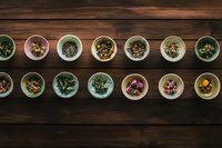 Small ceramic bowls with various dried herbs, flowers, and tea blends arranged in neat rows on dark wooden background, overhead view for wellness and tea ceremony.