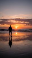 Silhouetted figure standing on wet beach sand at golden sunset, watching ocean waves and dramatic sky with warm orange and purple tones.
