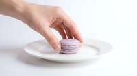 Hand placing a delicate lavender macaron on a white ceramic plate against a light background, evoking elegance and sweet indulgence.