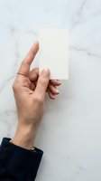 Female hand holding blank white card against marble background, wearing gold ring and dark sleeve. Mockup for business cards, invitations, or promotional materials.