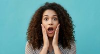 Surprised young African American woman with curly hair expressing shock and amazement with hands on cheeks against turquoise background for marketing campaigns.