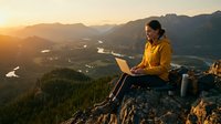Remote worker using laptop on mountain cliff at sunset overlooking valley with river and forest landscape below, freelance professional working outdoors.