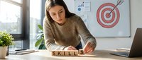 Young woman with dark hair and South Asian ethnicity, wearing a beige knit sweater, arranging wooden letter blocks spelling "skills" at her desk. Modern office setting with laptop, target board, charts, and plants visible. Concept for professional development, training, and career growth.