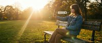 Young Caucasian woman sitting on park bench at golden sunset, holding phone with subscribe button, autumn trees glowing in warm sunlight.