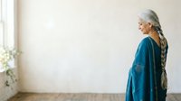 Senior woman with long gray braided hair wearing teal cardigan standing in bright minimalist interior space with window light, contemplative pose for lifestyle and wellness content.