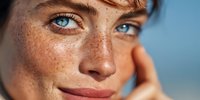 Close-up portrait of young woman with freckles and striking blue eyes against blurred blue background, conveying natural beauty and confidence.