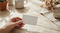 Hand holding blank white business card mockup on rustic wooden table with coffee mug and dried flowers for branding design presentation.