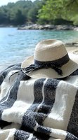 Straw sun hat with black ribbon on striped beach towel against blurred lake and forest background, summer vacation lifestyle setting for travel content.
