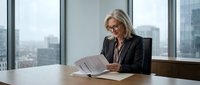 Professional mature blonde businesswoman reading documents at office desk with city skyline view through windows for corporate presentations.