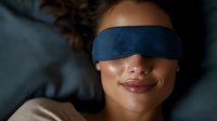 Young woman with curly hair wearing blue sleep mask lying in bed for restful night sleep and relaxation wellness routine.