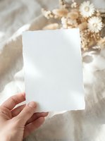 Blank white paper card mockup held in female hand with dried flowers on neutral linen background for wedding invitations and stationery design.
