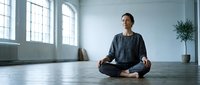 Young Caucasian man practicing meditation in lotus position on wooden floor in bright modern loft studio with large windows and minimalist interior design.
