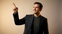 Confident young Caucasian businessman in dark suit pointing upward with finger, smiling at camera against neutral background for corporate presentations.