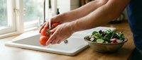 Male hands slicing fresh tomatoes on cutting board near bowl of green salad, preparing healthy meal in bright kitchen with natural window light.