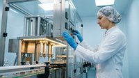 Scientist in white lab coat and hair net operating pharmaceutical manufacturing equipment in sterile facility, quality control testing.
