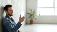 Young adult male with dark hair and beard, wearing casual denim shirt, holding smartphone with blank white screen in bright modern interior space for app mockup and technology marketing.