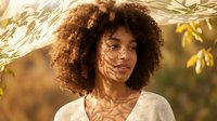Young African American woman with curly afro hair standing under tree shadows in warm golden sunlight wearing casual white top with natural makeup.