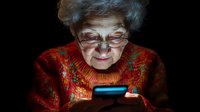 Elderly Caucasian woman with gray hair and glasses using smartphone in dark room, face illuminated by blue screen light wearing red sweater.