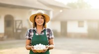 Farmer woman holding fresh eggs in basket at farm building, wearing straw hat and green apron, agricultural worker portrait.