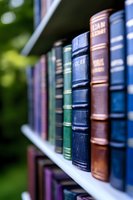 Row of vintage leather-bound books on white shelf against blurred green background, featuring blue, brown and green spines with gold lettering, shallow depth of field.