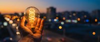 Hand holding glowing vintage Edison bulb against blurred city lights at sunset. Creative innovation and bright ideas concept for business and inspiration.