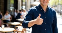 Young businessman giving thumbs up gesture at outdoor cafe terrace, expressing success and positive attitude in urban business environment.