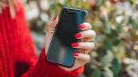 Female hand with red manicure holding smartphone against blurred green foliage background, wearing bright red knitted sweater.