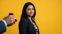 Professional South Asian businesswoman in black blazer smiling confidently at camera with male colleague using smartphone in background on vibrant yellow wall.