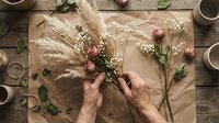 Florist arranging dried pampas grass, baby's breath, and pink carnations on kraft paper with natural twine and greenery on wooden work surface.