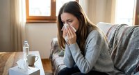 Young woman with long brown hair blowing nose into tissue while sitting on couch at home, feeling unwell with cold or flu symptoms in cozy living room.