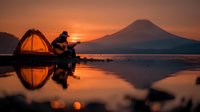 Camping musician playing guitar by lake at sunset with mountain silhouette reflected in calm water creating peaceful outdoor adventure scene.