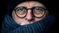 Close up portrait of mature Caucasian man wearing glasses, black knit hat and blue scarf wrapped around face, showing intense blue eyes against dark background.