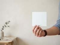 Close-up of male hand holding blank white card or paper against neutral beige wall background with minimal decor for mockup and branding purposes.