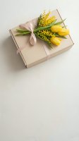 Elegant kraft gift box with pink ribbon, yellow tulips and mimosa flowers on neutral background for spring celebrations and special occasions.