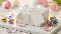 White gift box with satin ribbon surrounded by colorful Easter eggs and pink flowers on wooden surface for spring holiday celebration and gifting.