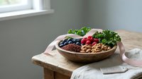 Wooden bowl filled with fresh berries, nuts, and leafy greens arranged on rustic table near window, perfect for healthy eating and wellness content.