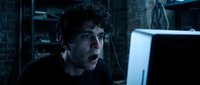Shocked young man with dark hair and olive complexion staring at computer screen in dimly lit tech environment, expressing surprise or concern while working late at night.