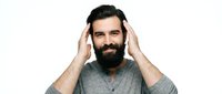 Bearded man with dark hair and facial hair wearing gray v-neck shirt, hands touching head, isolated on white background for portrait and lifestyle use.