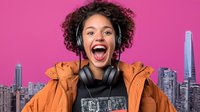 Excited young African American woman with curly hair wearing orange winter jacket and headphones laughing against pink background with city skyline.