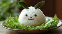 Cute bunny-shaped rice ball with mint leaves as ears, served on fresh lettuce. Creative Asian food art brings playful character to meal presentation.