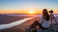 Young woman photographer with camera on tripod capturing golden sunrise over mountain lake valley from rocky summit during outdoor adventure.