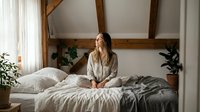 Young woman sitting peacefully on bed in cozy bedroom with exposed wooden beams and plants creating serene morning atmosphere for wellness lifestyle.