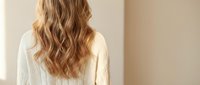 Young blonde woman with long wavy hair wearing white sweater standing with back turned in minimalist beige interior space for lifestyle content.