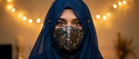 Middle Eastern woman wearing ornate golden face mask and blue hijab with decorative lights creating warm atmospheric background for cultural fashion portrait.